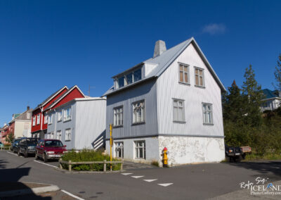 A street view featuring a mix of colorful buildings, including a prominent grey house with a peaked roof and multiple windows. The house is situated at a corner, with a white fence and a fire hydrant nearby. Several cars are parked along the side of the road, and the sky is clear and blue, adding to the vibrant atmosphere of the scene. Lush greenery can be seen in the background, completing the picturesque setting.
