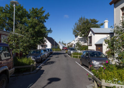 A quiet street scene featuring parked cars along the sides, with trees and shrubs providing greenery. Residential buildings line the street, some with traditional architectural elements. The sky is clear and blue, suggesting a sunny day. The perspective shows the street leading into the distance, creating a sense of depth.