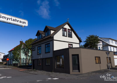 A street view showcasing a modern, two-story building with a combination of white siding and dark trim. The house features several large windows with red accents and a distinctive angular roof. In the foreground, a stop sign and a street sign that reads "Smyrlahraun" are visible, along with a cobblestone street and a nearby green building with a red roof. The sky is clear and blue, creating a bright atmosphere.