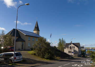 A view of a charming church with a tall spire and cross on top, situated on a hillside. The church features arched windows and is painted a light color. In front of the church, there are parked cars and a bicycle. To the right, another building with a dark roof is visible. The scene is set under a bright blue sky with scattered clouds, and there are a few trees around, adding greenery to the landscape. A road runs along the lower section of the image, indicating a residential area nearby.