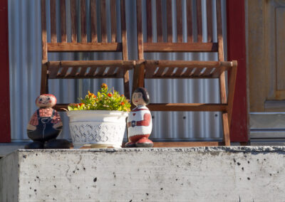A sunlit outdoor scene featuring two wooden rocking chairs placed on a concrete step. Between the chairs, there is a white potted plant with colorful flowers. Flanking the pot are two decorative dolls, one dressed in a checkered shirt and overalls, and the other in a red and white outfit, both adding a whimsical touch to the setting. In the background, there is a corrugated metal wall and a wooden door with a decorative glass panel.