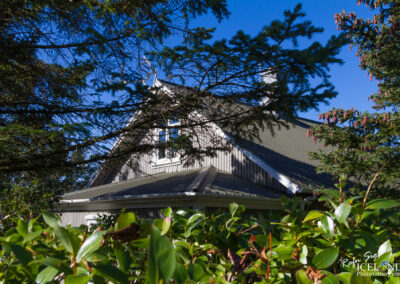 A roof of a house is partially visible, framed by green foliage and branches. The house has a sloped roof, white trimming, and a few windows that reflect the sunny blue sky above. The foreground is filled with lush greenery, while the background shows clear blue skies with a few scattered clouds.