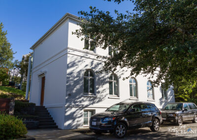 A modern white building with arched windows is seen in this image. It has a wooden door at the front and is surrounded by greenery, including trees and shrubs. Two vehicles, one dark SUV and another larger vehicle, are parked in front of the building. The sky is clear and blue, providing a bright backdrop to the scene. The ground features stone steps leading up to the entrance.