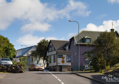 A residential street with sloped pavement featuring two houses on the right side. The first house has a dark roof and a mix of white and wooden siding, while the second house is painted purple with a green roof. There are a couple of parked cars, one silver and one orange, and a street sign indicating a left turn. Lush greenery and trees are present on the left side, under a clear blue sky with a few clouds.