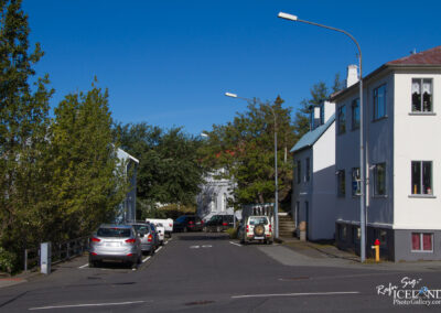 A quiet street view featuring parked cars along both sides of the road, bordered by trees and modest white buildings. The sky is clear and blue, and street lights are visible on the left. In the distance, a glimpse of another road can be seen, leading to a building with steps and lush greenery around.