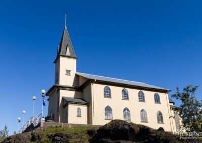 A light-colored church building with a tall spire and a cross on top is set against a clear blue sky. The structure features multiple arched windows and has a sloped roof. In the foreground, there are decorative lampposts and a rocky landscape with some greenery visible.