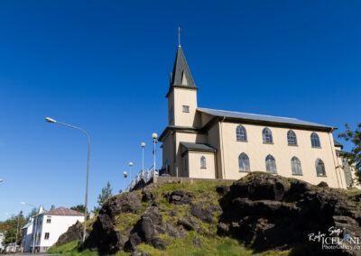 A tall church with a pointed steeple sits on a rocky hillside, surrounded by a bright blue sky. The church features large arched windows and is constructed with light-colored materials. In the foreground, there are patches of grass and rocks, while streetlights and a few trees are visible nearby. Nearby buildings have white exteriors, adding to the scenic view.