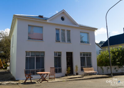 A modern white house with large windows, featuring a sloped roof and a circular window at the peak. In front of the house, there is a small wooden table surrounded by two chairs and a long bench, providing a cozy outdoor seating area. The setting includes well-maintained greenery and a clear blue sky in the background.