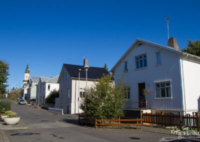 A tranquil street scene featuring a row of colorful houses on a clear sunny day. The houses are primarily two stories tall with varying architectural styles, including a white wooden house on the right and a building with a dark roof on the left. A church spire can be seen in the distance, alongside trees and shrubs lining the street. The area is well-maintained, with a planted flower bed and a wooden fence around a small garden area in the foreground. The bright blue sky adds to the overall serene atmosphere.