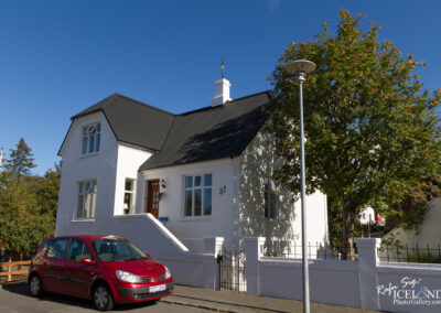 A charming white house with a black roof sits by the side of the street. The house features large windows and a wooden front door, with the number "37" displayed prominently on the facade. In front, a red car is parked, and a well-maintained garden with a tree can be seen nearby. The sky is clear and blue, adding to the inviting atmosphere of the scene.