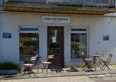 A quaint storefront of a cafe or shop named "Gamla Matarbúðinn" with a white exterior and a brown door. There are large windows displaying products inside, and a small round table with four wooden chairs set outside on a paved area. A piece of paper is taped to the window, and there is a photo displayed on the wall next to the entrance. The area is surrounded by grass and small plants.