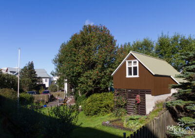A sunny urban scene featuring a quaint brown house with a green roof and white window trim. The house is surrounded by lush greenery, including trees and bushes, as well as a well-kept garden. In the background, there are other buildings and a clear blue sky. A couple of people can be seen enjoying the outdoors, while a flagpole stands to the left of the house.