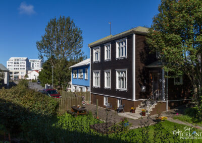 A picturesque street scene featuring a charming wooden house painted in dark brown with white trim, showcasing several large windows. Adjacent to it is a blue house with white accents. In the foreground, a small garden area with wooden picnic tables and potted flowers is visible, bordered by a wooden fence. Several vehicles are parked nearby, and tall trees provide shade against a clear blue sky.