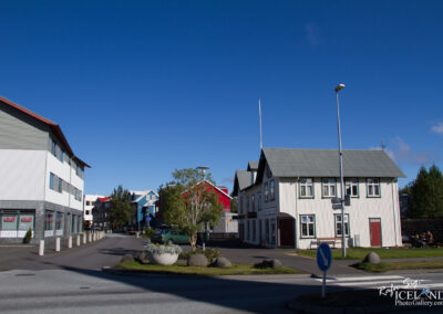 A street view featuring a mix of modern and traditional buildings. On the left, a contemporary building with gray and white panels and large windows, along with storefronts. On the right, a charming, two-story house with white siding and a green roof. In the background, colorful houses with red, blue, and green roofs can be seen. The scene is bright with a clear blue sky and a few scattered clouds. There are trees and greenery in the area, and a roundabout is visible in the foreground.