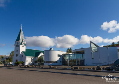 The image showcases a modern building adjacent to a traditional white church with a green steeple. The church features a clock and a simple architectural design, while the modern building has smooth curves and large glass windows. The scene is set against a blue sky dotted with fluffy clouds, and there are stone formations and greenery in the foreground, adding to the peaceful ambiance of the location.