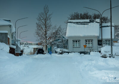 A snowy street scene featuring residential houses blanketed in snow. The foreground shows a snow-covered road with tire tracks leading deeper into the neighborhood. Streetlights line the sides, and a few trees are visible, some also covered in snow. In the background, additional houses and a hint of a distant hillside can be seen, under a gray sky with a subtle glow. A car is parked on the roadside, partially buried in snow, and a street sign is visible nearby.