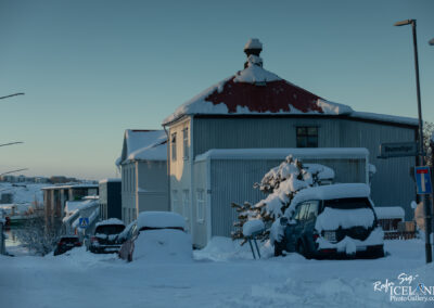 A snowy street scene featuring several buildings with snow-covered roofs. One building has a distinct red roof and the others are painted in lighter colors. Parked vehicles are mostly obscured by thick snow, and a tree is laden with snow nearby. Street signs and lampposts are visible, indicating that the area is urban. The sky is clear, reflecting a serene winter atmosphere.