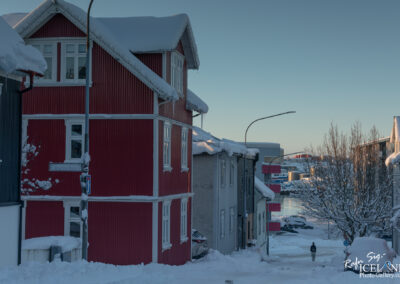 A quiet winter scene featuring snow-covered streets lined with colorful houses. Prominent in the foreground is a red building with white trim, neighboring a dark blue structure. Snow blankets the rooftops and coats the ground, while a streetlight and a traffic light stand amid the winter landscape. In the background, the street leads toward a body of water, hinting at a scenic area. A solitary person walks down the road, adding a sense of tranquility to the snowy environment.