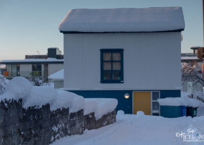 A quaint, two-story house covered in a thick layer of snow. The house features a blue lower section and white upper siding, topped with a snow-laden roof. A bright yellow door stands at the front, with a round light beside it. Snow blankets the pathway leading to the entrance, and a stone wall partially obscures the view. In the background, modern buildings can be seen, also dusted with snow, under a clear sky.