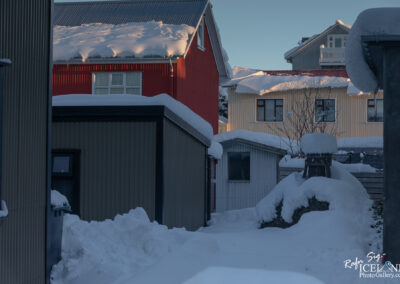 A snowy scene depicting a narrow pathway between buildings, with varying colored rooftops covered in white snow. The background features a red building with a white window, and neighboring structures in shades of gray and yellow, all accented by the soft morning light illuminating the snow. The ground is heavily blanketed in snow, creating a tranquil winter atmosphere.