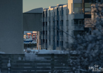 A view through a narrow opening between two buildings, showcasing a marina in the background. The reflection of light glimmers on the water, highlighting a blue boat and red structures. The scene is set in a winter environment, with snow covering the rooftops and surrounding areas, creating a calm and serene atmosphere.