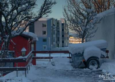 A snowy pathway leads through a residential area. On the left, a red building with a lamp post is partially visible. In the foreground, a trailer covered in snow sits at the edge of the path. In the background, a modern multi-story building with a blue and gray facade is seen. Bare branches of trees are visible in the scene, contrasting with the white snow and a clear blue sky.