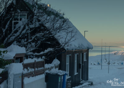 A cozy, snow-covered house is partially visible, tucked against a backdrop of a serene winter landscape. The roof is thick with snow, and nearby trees are bare with snow clinging to their branches. In the foreground, a wooden fence surrounds the property, and blue trash bins sit near the house. In the distance, a calm body of water reflects soft light, with a snowy path lined by street signs leading toward it. The overall atmosphere is tranquil and picturesque, capturing the essence of a winter day.