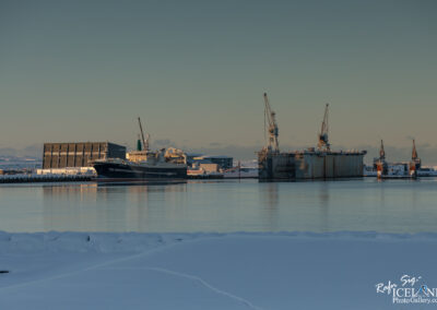 A tranquil harbor scene featuring a large boat docked at a pier, with a large industrial structure in the background. Two prominent cranes are visible near the water, along with another large vessel nearby. The landscape is covered in smooth, white snow, and the water reflects the soft colors of the sky, suggesting a calm atmosphere.