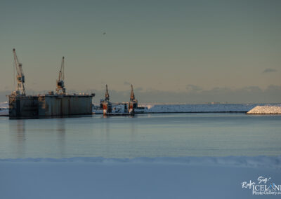 A tranquil winter scene featuring a calm body of water reflecting the soft light of the sky. In the foreground, there is a snow-covered bank leading to the water. In the background, two large, industrial vessels are docked, with cranes positioned above them. A long, snow-covered pier extends to the right, while low clouds and a soft gradient in the sky create a serene atmosphere. The overall mood of the image is peaceful and stark, highlighting the contrast between the industrial structures and the natural environment.