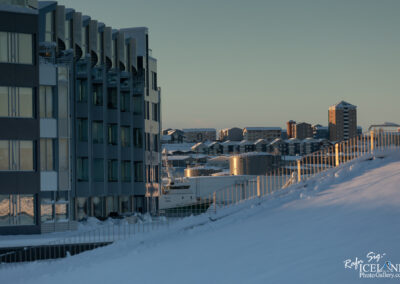 A winter landscape featuring modern buildings with large glass windows and snow-covered ground. In the foreground, a gently sloping hill is blanketed in white snow. On the left, a contemporary multi-story building stands, showcasing a mix of gray and reflective surfaces. In the background, additional buildings of varying heights and designs sit atop a snowy hill, with hints of sunlight illuminating their rooftops. A white boat is partially visible in the harbor, among silos and other structures, contributing to the overall serene winter atmosphere.