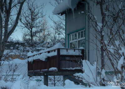 A wooden balcony covered in snow extends from a building, surrounded by trees also blanketed in white. The building features large windows and a metal exterior, with icicles hanging from the roof. The scene captures a tranquil winter atmosphere, emphasizing the abundance of snow and the natural surroundings.