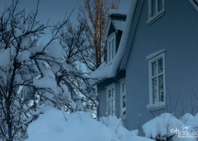 A close-up view of a blue, snow-covered house surrounded by bare trees and thick layers of snow. The roof has snow piled on top, and icicles hang from the eaves. Sunlight casts a warm glow on part of the house, contrasting with the cool, wintry scene. The background features a clear, dark blue sky.