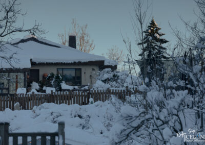 A cozy house is blanketed in snow, with a thick layer covering the roof and tree branches nearby. A wooden fence surrounds the yard, which is also covered in snow. Scattered trees, some with snow on their branches, are visible in the background, creating a serene winter landscape under a clear sky.