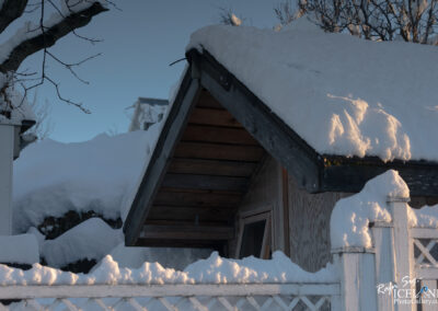 A snowy landscape featuring the corner of a building with a steeply sloped roof covered in fresh snow. The roof has a wooden overhang and is partially illuminated by soft sunlight, creating a warm glow against the cold white snow. In the background, snow-laden trees can be seen against a clear blue sky. A white picket fence at the bottom adds to the wintery scene.