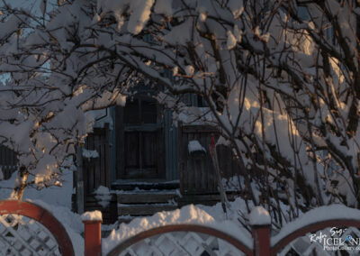 A snowy scene featuring a wooden house partially obscured by snow-laden branches. The entrance to the house is visible, with a set of steps leading up to a slightly open door. The foreground includes a red wooden fence, and the ground is covered in a thick layer of snow, creating a serene winter atmosphere with soft sunlight illuminating the scene.