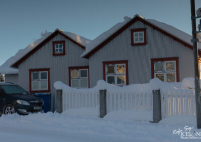 A snow-covered residential house with a distinctive tin exterior and red trim. The building features large windows with snow resting on the roof and surrounding fence. A parked dark-colored car is visible in front of the house, partially obscured by snow. The scene is set against a clear blue sky, suggesting a cold winter day.