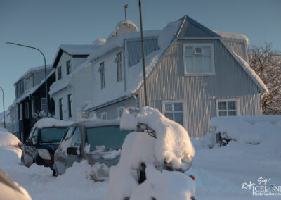 A snow-covered street scene featuring several houses, with one large building showing a combination of white and gray siding, and a roof topped with thick snow. The foreground includes parked cars and a motorcycle, all heavily blanketed in snow. The sky is clear with soft sunlight illuminating the scene.
