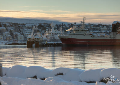A tranquil harbor scene featuring several fishing boats docked in the water, surrounded by snow-covered landscapes and houses in the background. The surface of the water reflects the calmness of the scene, while snow drifts sit along the foreground. The sky is painted with soft hues of orange and blue as the sun rises or sets, adding a warm glow to the cold winter atmosphere.