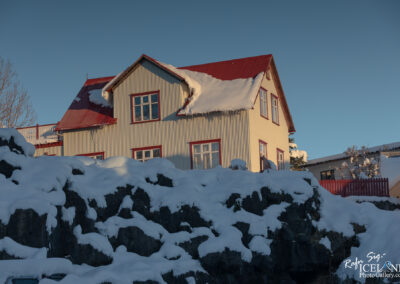 A snow-covered landscape featuring a quaint house with a red roof and white walls, framed by large rocks draped in snow. The house has multiple windows, some of which reflect the clear blue sky. In the background, additional structures are partially visible, also blanketed in snow. The scene is lit by soft sunlight, casting gentle shadows across the snowy terrain.