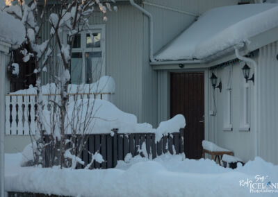A snow-covered exterior view of a house with a dark wooden door, surrounded by white snow and a few snow-laden trees. There is a railing at the porch, and some holiday lights are visible on the side of the house. The overall scene conveys a tranquil winter atmosphere.