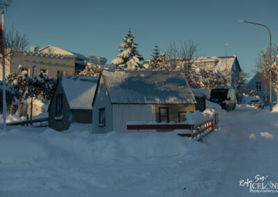 A snow-covered street lined with small houses, some with metal roofs partially buried in snow. Trees dusted with snow are visible, and the bright blue sky contrasts with the white landscape. A parked van is seen on the right, and the road is flanked by snowbanks, creating a peaceful winter scene.