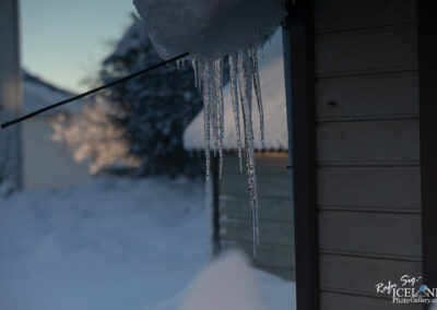 A close-up view of icicles hanging from a roof edge, with a blurred background of a snow-covered landscape and a soft sunset glow. The scene captures a cold winter atmosphere, focusing on the delicate structures of the icicles against a wooden surface.