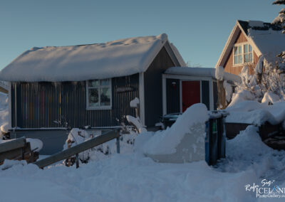 A cozy, snow-covered house with a sloped roof blanketed in thick snow sits against a clear blue sky. The house has vertical wooden siding and a bright red door. Snow drapes over the front railing and bushes, while a few icicles hang from the roof edge. In the foreground, a snow-encased pathway leads to the house, alongside two colorful garbage bins partially obscured by snow. Additional houses can be seen in the background, enhancing the winter landscape.