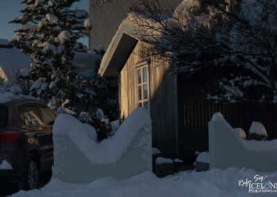 A snow-covered scene featuring a house with a wooden exterior and large windows, illuminated by the soft light of the setting sun. A dark-colored car is parked nearby, partially obscured by snow. In the foreground, there is a white, snow-covered stone wall with decorative edges, and snow-laden trees and shrubs can be seen in the background, creating a serene winter atmosphere.