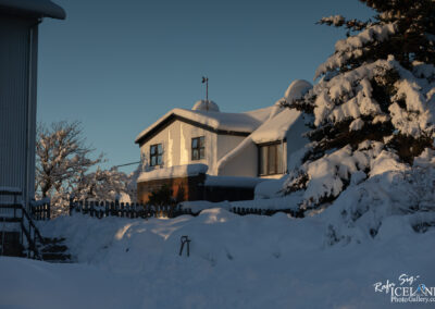 A snow-covered house with a peaked roof is surrounded by trees laden with heavy snow. The scene is illuminated by warm morning light, contrasting with the clear blue sky. A wooden fence encloses a small pathway leading to the house, while stairs are partially visible on the left, covered in snow. The overall atmosphere conveys a peaceful winter landscape.