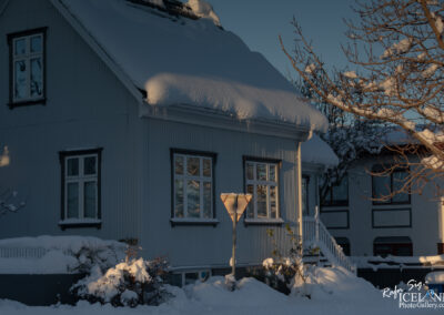 A snowy scene featuring a two-story house with a light blue exterior. The roof is heavily laden with snow, and icicles hang from the overhang. In the foreground, a snow-covered bush is illuminated by the low sunlight, and a stop sign partially buried in snow is visible. Nearby, additional buildings can be seen, also blanketed in snow, under a clear sky.