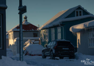 A street scene in a snowy landscape featuring several houses. The house in the foreground is painted blue with white trim, and icicles hang from its eaves. Next to it, a red-roofed house is partially obscured by snow. Two cars, one covered in snow and the other parked, are visible on the street. The sun casts a warm golden light on the houses, contrasting with the cold blue tones of the snow. A street sign is partially visible in the foreground.