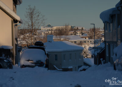 A snow-covered residential street in a quiet neighborhood, with houses partially buried in snow. A layer of snow rests on the roofs of the buildings and cars parked along the street. In the background, more houses and trees are visible, indicating a tranquil winter scene. The sky is clear, suggesting a bright and sunny day despite the cold.