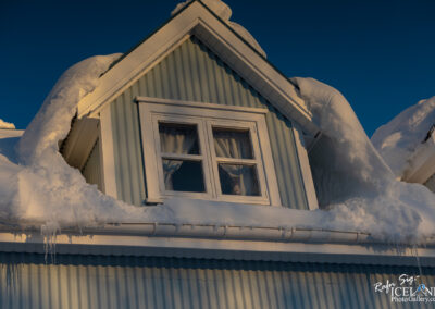 A close-up view of a snow-covered roof of a house, featuring a textured blue wall and white-trimmed windows. Snow is piled high on the roof edges, creating soft, rounded shapes. Icicles are hanging from the roof's eaves, and the scene is illuminated by warm sunlight against a clear blue sky.