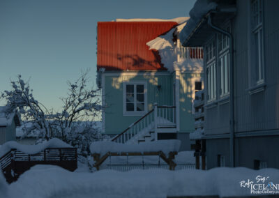 A winter scene featuring two houses partially covered in snow, with one house displaying a bright red roof and the other painted in a light green hue. A snow-laden tree with branches is visible in the foreground, and a staircase leading to a balcony is also seen. The light casts soft shadows across the scene, highlighting the textures of the snow and buildings against a clear blue sky.